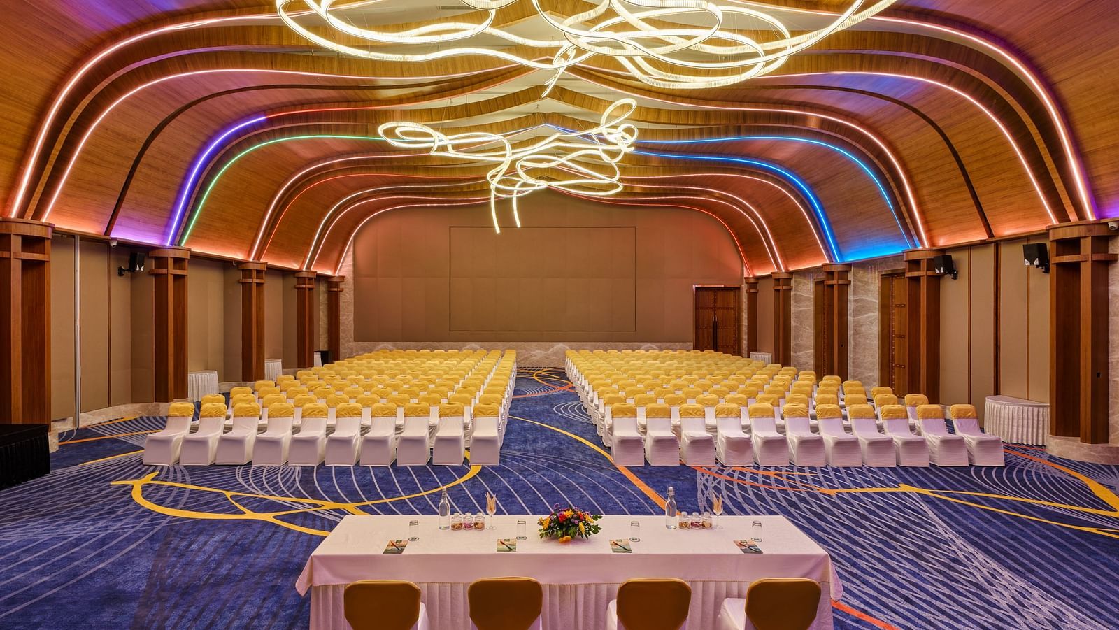 Image of the meeting hall Boito Voyage at Swosti Premium Beach Resorts in Puri featuring a large sitting area for audience under warm lighting and four chairs arranged with a large table facing the audience