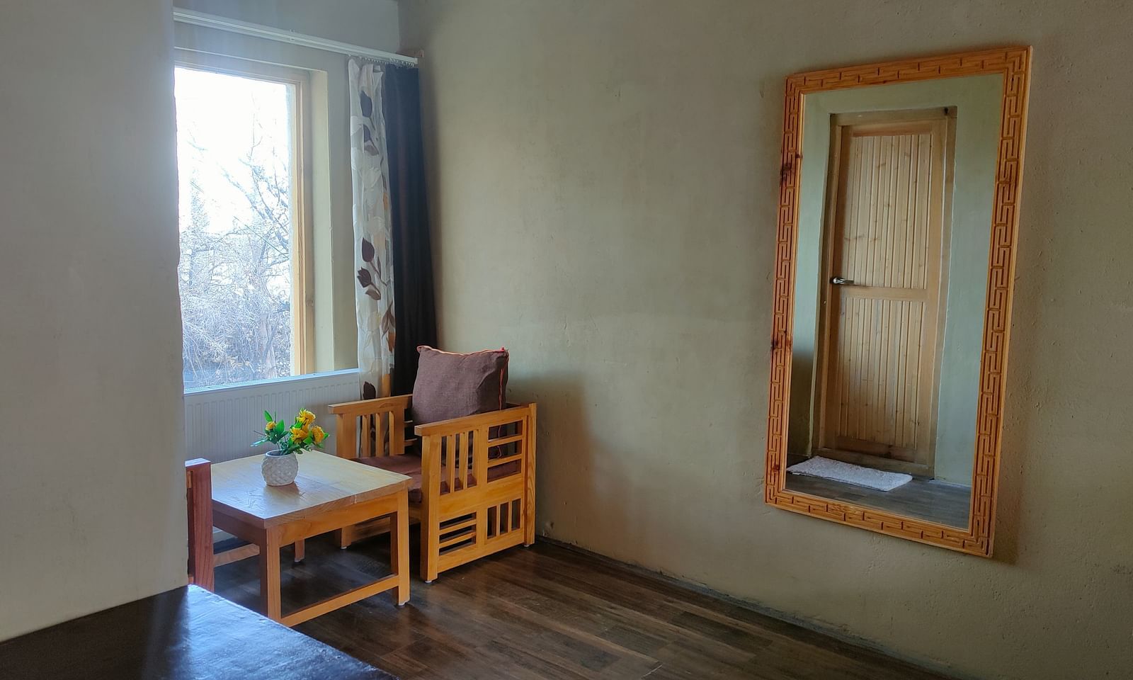 A minimalist corner of Deluxe Cottage is captured, featuring a wooden-framed mirror, a small table with a lamp, and a window offering natural light, complemented by a wooden stool at White Sands Resort, Ladakh.