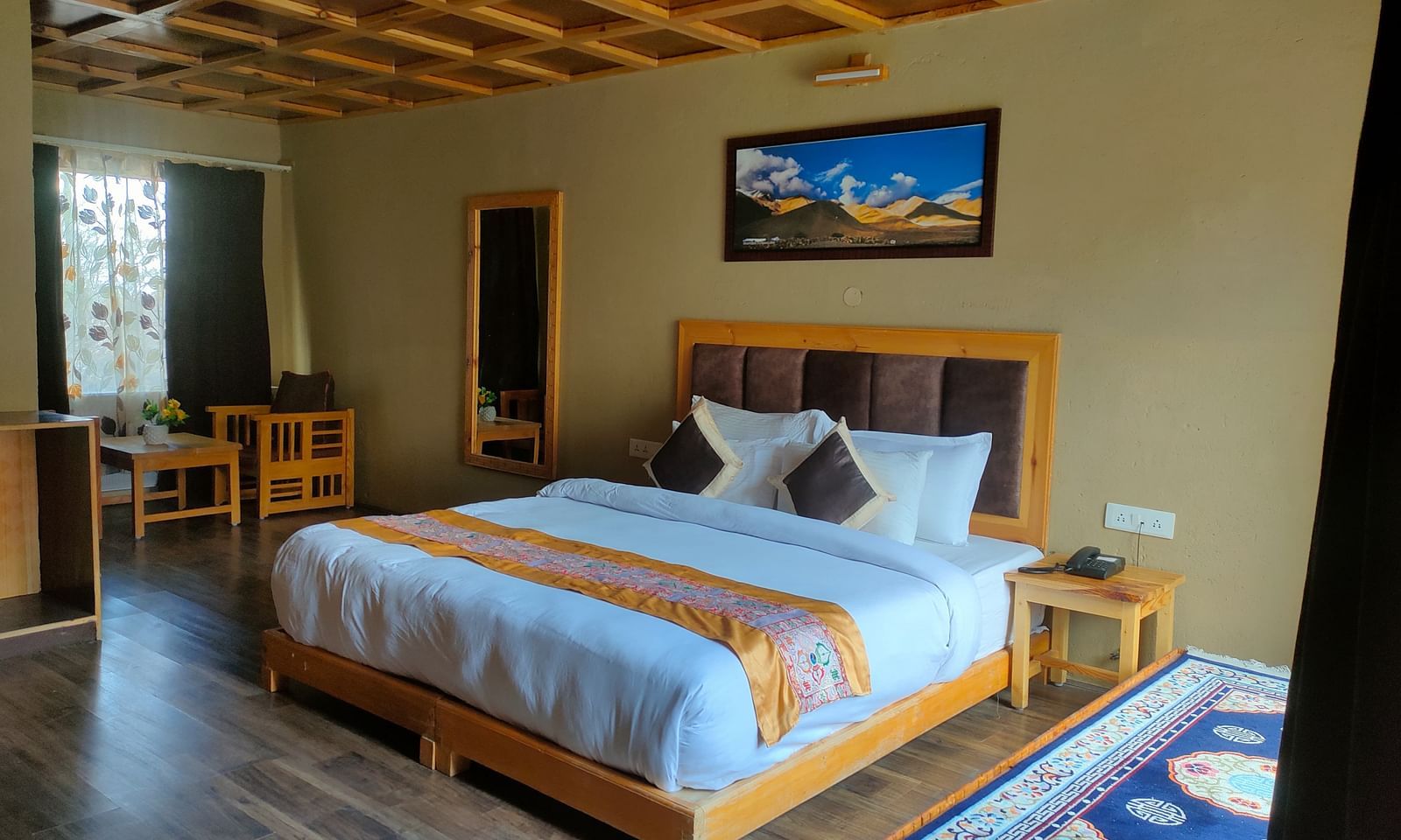 Deluxe Cottage showcases a neatly made king size bed with a striped throw, accompanied by wooden furniture, a decorative ceiling, and a framed picture at White Sands Resort, Ladakh.
