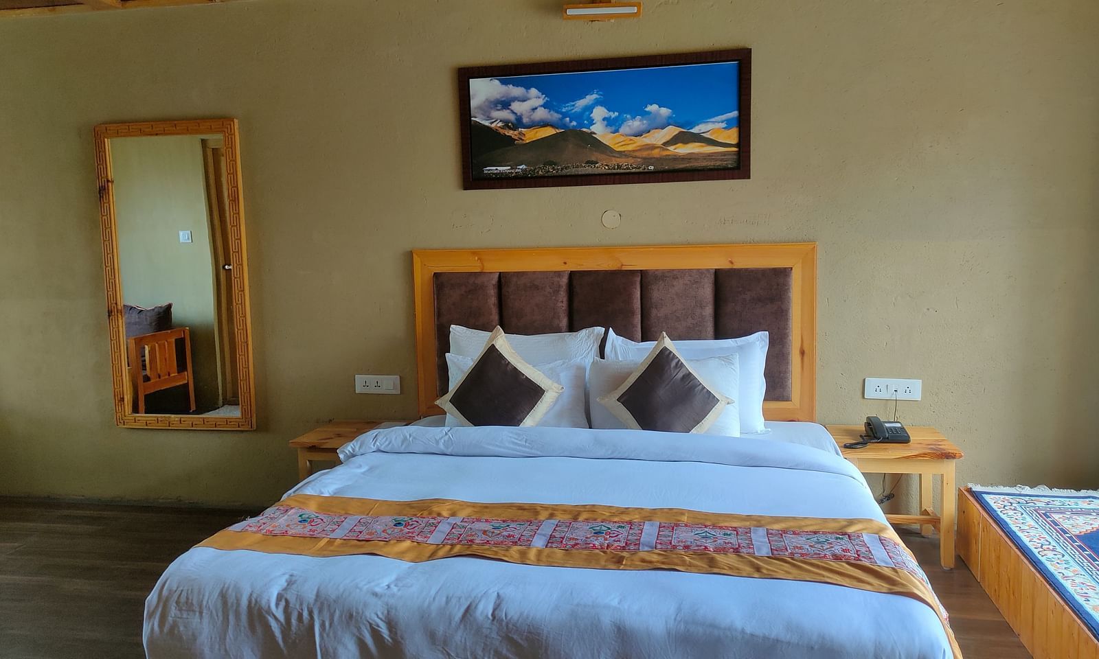 A well-lit Deluxe Cottage features a king size bed with white linens and a patterned throw, flanked by a wooden headboard and bedside table, with a full-length mirror and a framed picture at White Sands Resort, Ladakh.