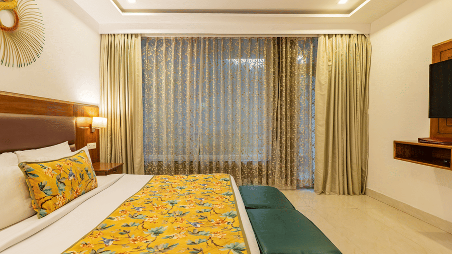 A bedroom with a neatly made bed, 2 vanity stools, a decorative ceiling, a tiled floor, and sheer curtains with floral patterns | Summit by the Ganges Beach Resort & Spa, Rishikesh