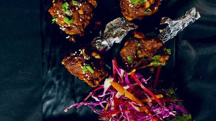 Black rectangular platter of Drums of Heaven chicken pieces with sesame seeds and spring onion, garnished with a slaw of red cabbage and carrot.