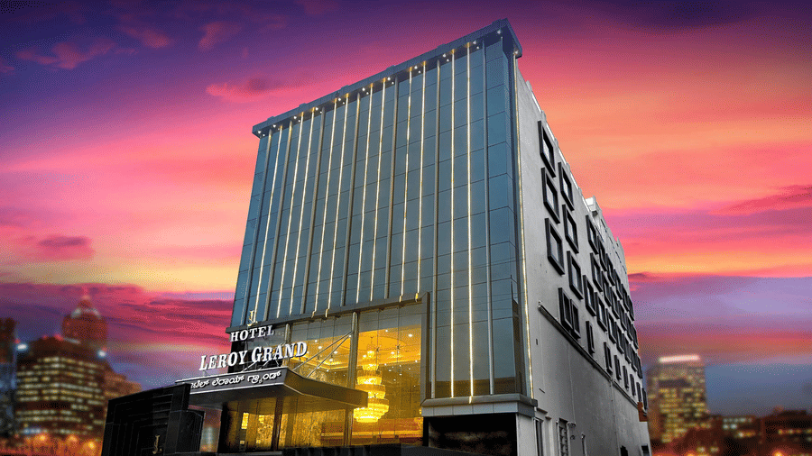 Facade of Leroy Grand, Yeshwanthpur, featuring a tall building with a brightly lit lobby area along with a beautiful sunset sky with pink and orange hues.