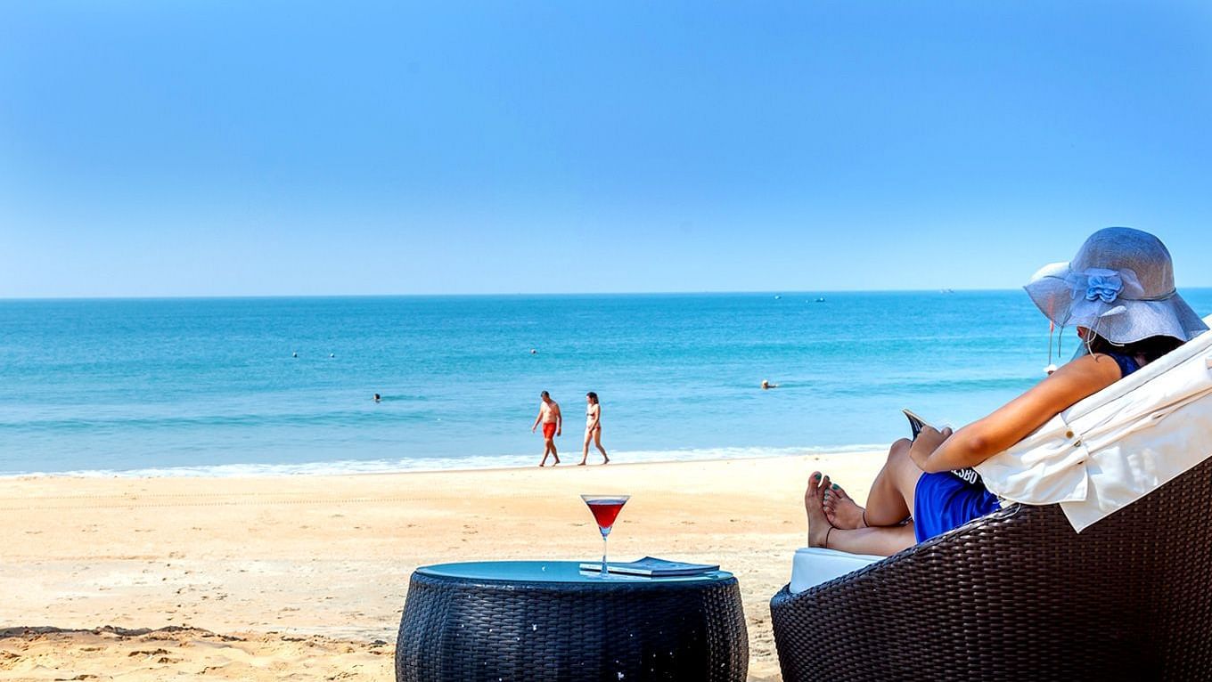 a woman sitting on a lounger with a drink on the table on the beach - Caravela Beach Resort Goa