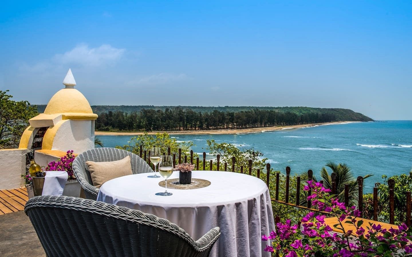 An outdoor dining table with white tablecloth and chairs, set on a patio overlooking a vast bay with blue water and distant land. A yellow dome or turret is visible on the left from Fort Tiracol, Goa.