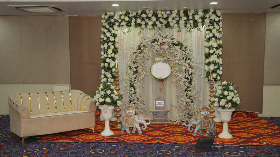 A decorated stage setup at Ambar Sarovar Portico, Gandhidham displays a patterned couch, floral arrangements, elephant figurines, and carpets.