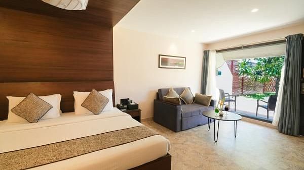 A well-lit hotel room features a king-size bed with neutral linens, a gray loveseat, a round glass table, and a balcony overlooking greenery - MGM Mark Whitefield
