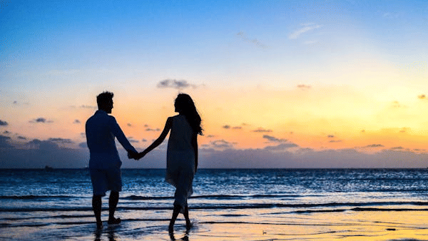 Two people hold hands facing the ocean at sunset, their silhouettes reflected on the wet sand.