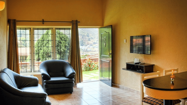 Living room with two black armchairs, a round table, wall-mounted television, and large sliding glass doors opening to a garden area at Green Pastures, Kodaikanal.
