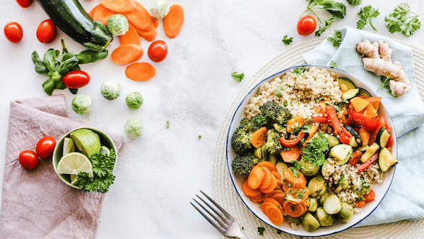 A colorful plate of healthy food with brown rice, sautéed vegetables, and herbs, surrounded by fresh ingredients on a white surface.