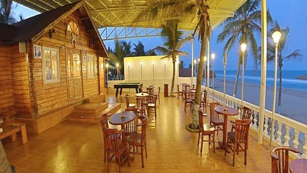 An open-air dining area with wooden tables, chairs, palm trees and light poles beside the sea at Hotel Mamallaa Heritage, Mahabalipuram.