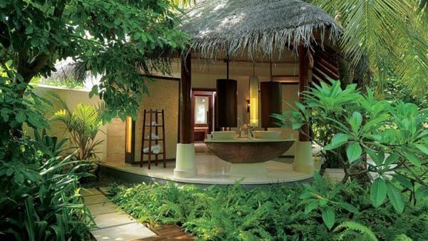 A small cottage with 2 wash basins, and a towel rack, surrounded by green plants and trees at Hotel Mamallaa Heritage, Mahabalipuram.