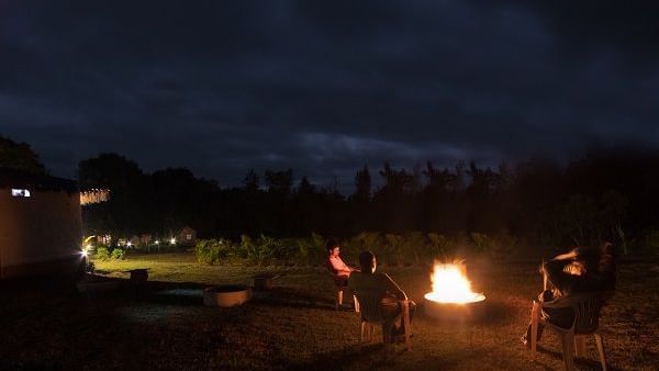People sitting around a campfire at night in an open area with faint lights and trees in the background.