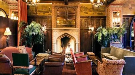 A seating area with 2 sofas, 5 sofa chairs, 2 tea tables, lamps and a fireplace at Hotel Mamallaa Heritage, Mahabalipuram.