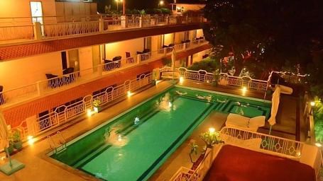 A swimming pool with a lounge at poolside and surrounded by room balconies and lights in the evening at Hotel Mamallaa Heritage, Mahabalipuram.
