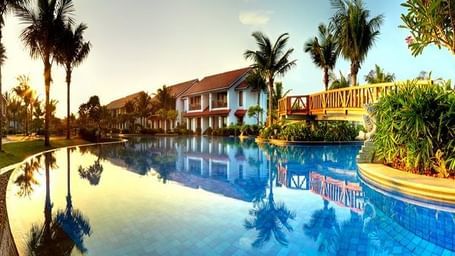 An outdoor pool with palm trees and a bridge with buildings in background at Hotel Mamallaa Heritage, Mahabalipuram.