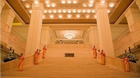 A wide staircase with railings, ceiling lights and hotel staff on either side of the stairs in the lobby area at Hotel Mamallaa Heritage, Mahabalipuram.