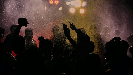  Silhouetted figures with raised hands at a lively, crowded event with bright, colourful stage lighting.