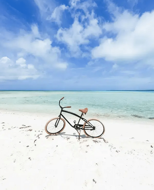 A bicycle parked on white sand near clear blue water under a partly cloudy sky, with no one around.