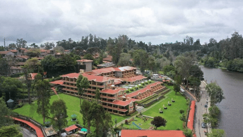 Facade of The Carlton in Kodaikanal and lawn outside the hotel. The location is surrounded by trees, a road on one side, and next to it there is a lake.