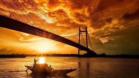A glowing sunset behind Howrah Bridge, as a traditional boat glides over the calm Hooghly River in Kolkata.