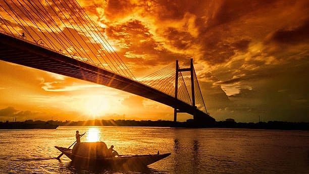 A glowing sunset behind Howrah Bridge, as a traditional boat glides over the calm Hooghly River in Kolkata.