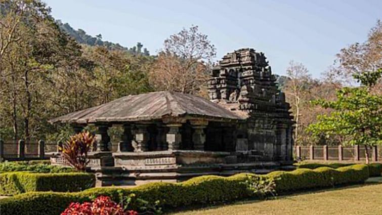  An ancient stone temple with intricate carvings and a multi-tiered roof, surrounded by a well-maintained lawn and manicured hedges, with lush green trees and hills in the background under a clear sky.