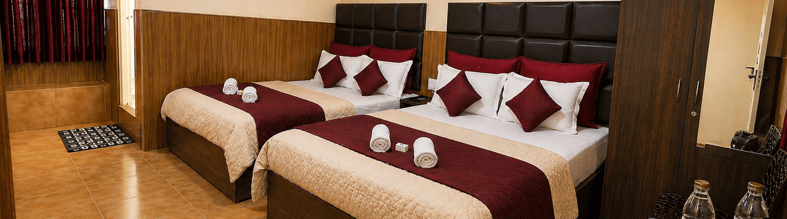 2 large beds arranged side by side with a wardrobe, a wall-mounted fan, and a kettle with water bottles on a table in the Deluxe Quadruple Room at BN Tourist Home, Ooty.