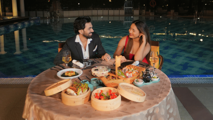 A man and a woman seated at a round table with multiple dishes placed beside a swimming pool at The Suryaa, New Delhi.