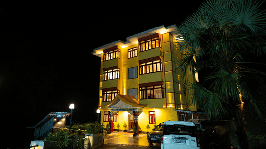 The well-lit facade of Voyage The Royal Retreat, featuring large windows, trees beside, and vehicles in front of the building at night.