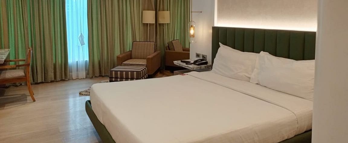 A warmly lit executive room in Mirage Hotel, featuring a king size bed, a lounge chair, a work desk, and a wall to wall window with partially open drapes.