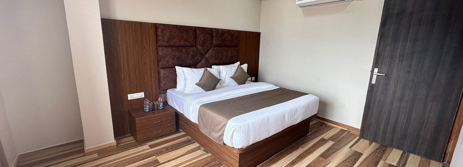 A bedroom includes a double bed with white and beige linens, dark wooden furniture, and a ceiling fan - Kankidham Lords Inn