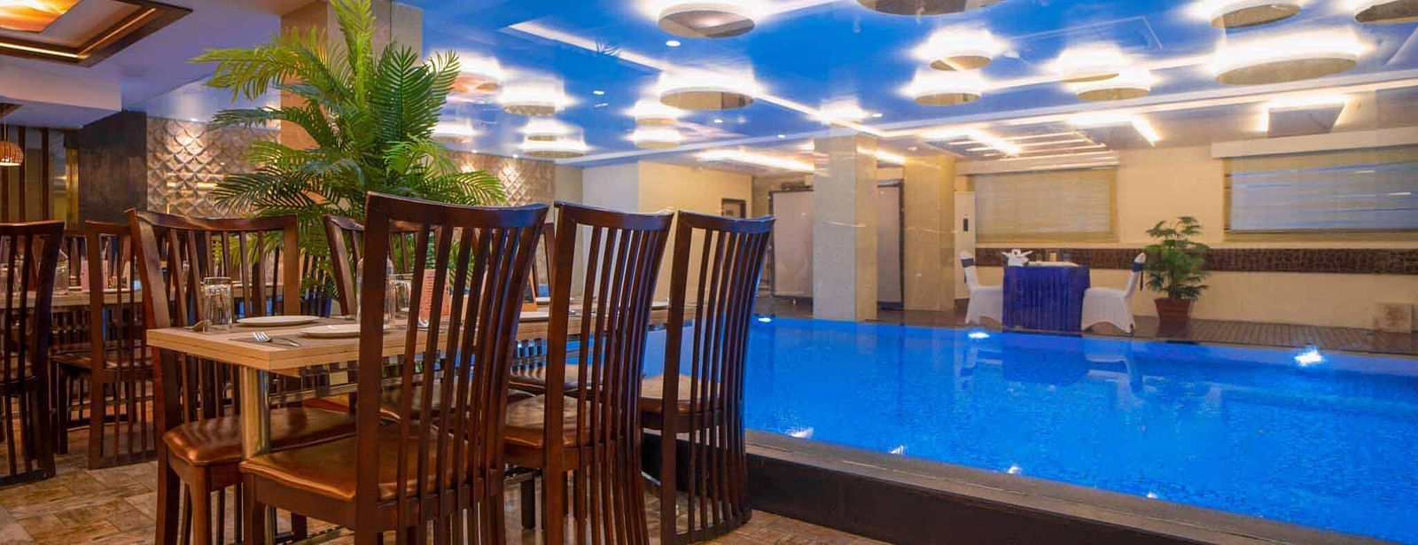 A dining area at Mansha Hotel Regency with a distinctive blue-lit ceiling, dark wooden tables and chairs, and a pool in the foreground.