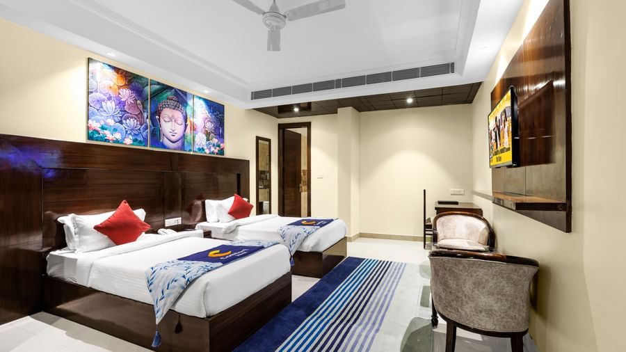 Elegant room with twin beds, ceiling fan, rug, chairs, wall-mounted TV, and colourful artwork above the beds at Inde Reva Resorts Rishhikesh.