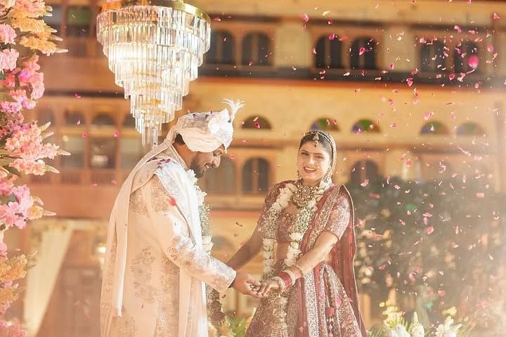 A couple stands under a  floral chandelier, exchanging smiles and holding hands as petals fall around them in celebration. - Umaid Palace - A Lakeside Heritage Palace Resort