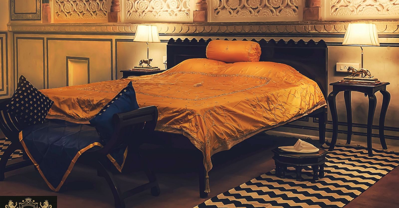 A bedroom at Umaid Vilas Jaipur with a bed dressed in golden fabric, two lamps, and decorative wall cutouts.