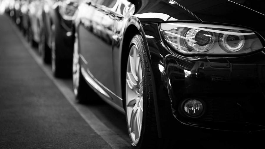 A row of parked cars, lined up along a road with a close-up focus on the front headlight of a black car.