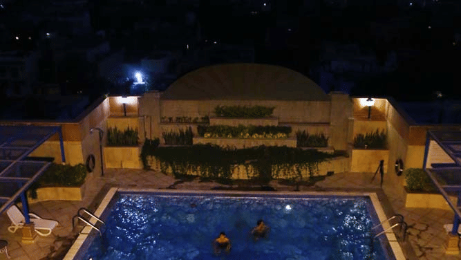 Rooftop swimming pool at The Bristol Hotel, Gurgaon at night with guests swimming and the illuminated city skyline visible in the background.