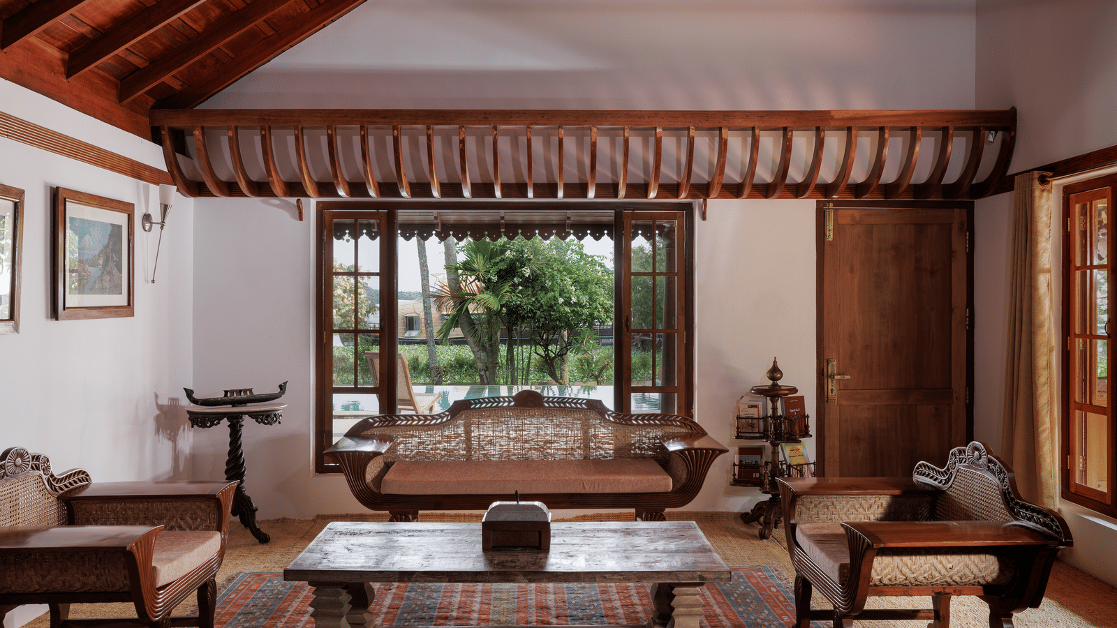 A shot of a room showcasing the sitting area with wooden couches, a coffee table, and a huge window overlooking the view - BluSalzz Villas - Muhamma Resort, Alleppey