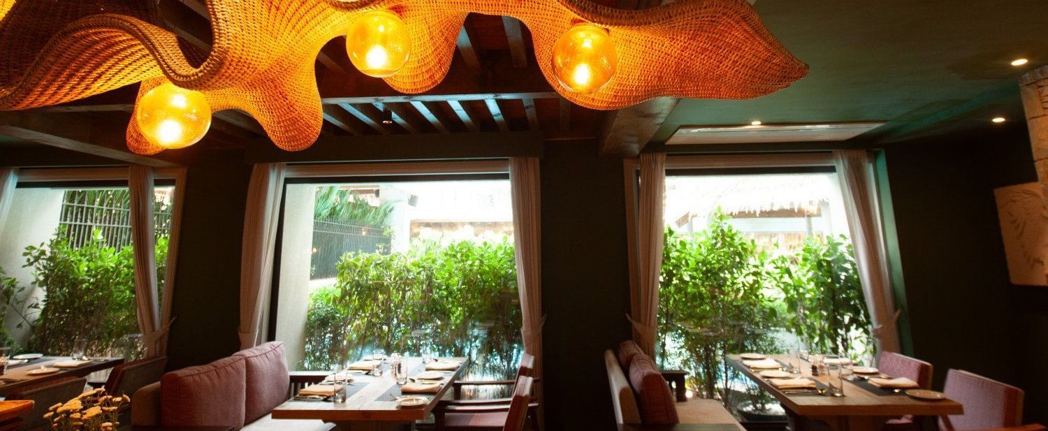 A detailed shot of the interior of Mila Restaurant at The Evren, Vagator, highlighting the unique, woven light fixtures hanging from the wooden ceiling. The restaurant has large windows with a view of greenery and comfortable seating.