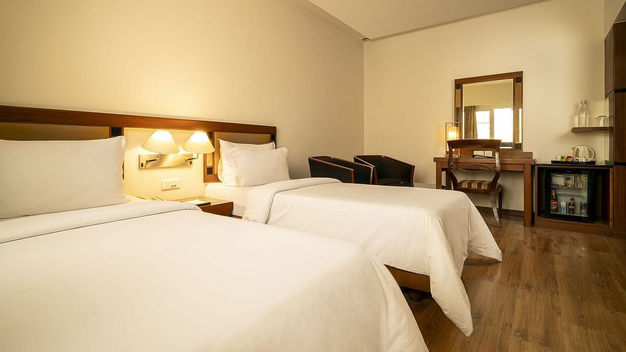 A warmly lit hotel room presents two single beds with white linens, a wooden desk with a mirror, and a mini-fridge at The Residency Karur.