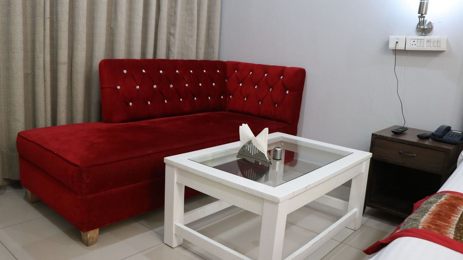 A cosy corner with a red tufted sofa, a white square table, and a bedside table, illuminated by a wall lamp against a soft gray wall. - Hotel Diamond Plaza, Chandigarh.