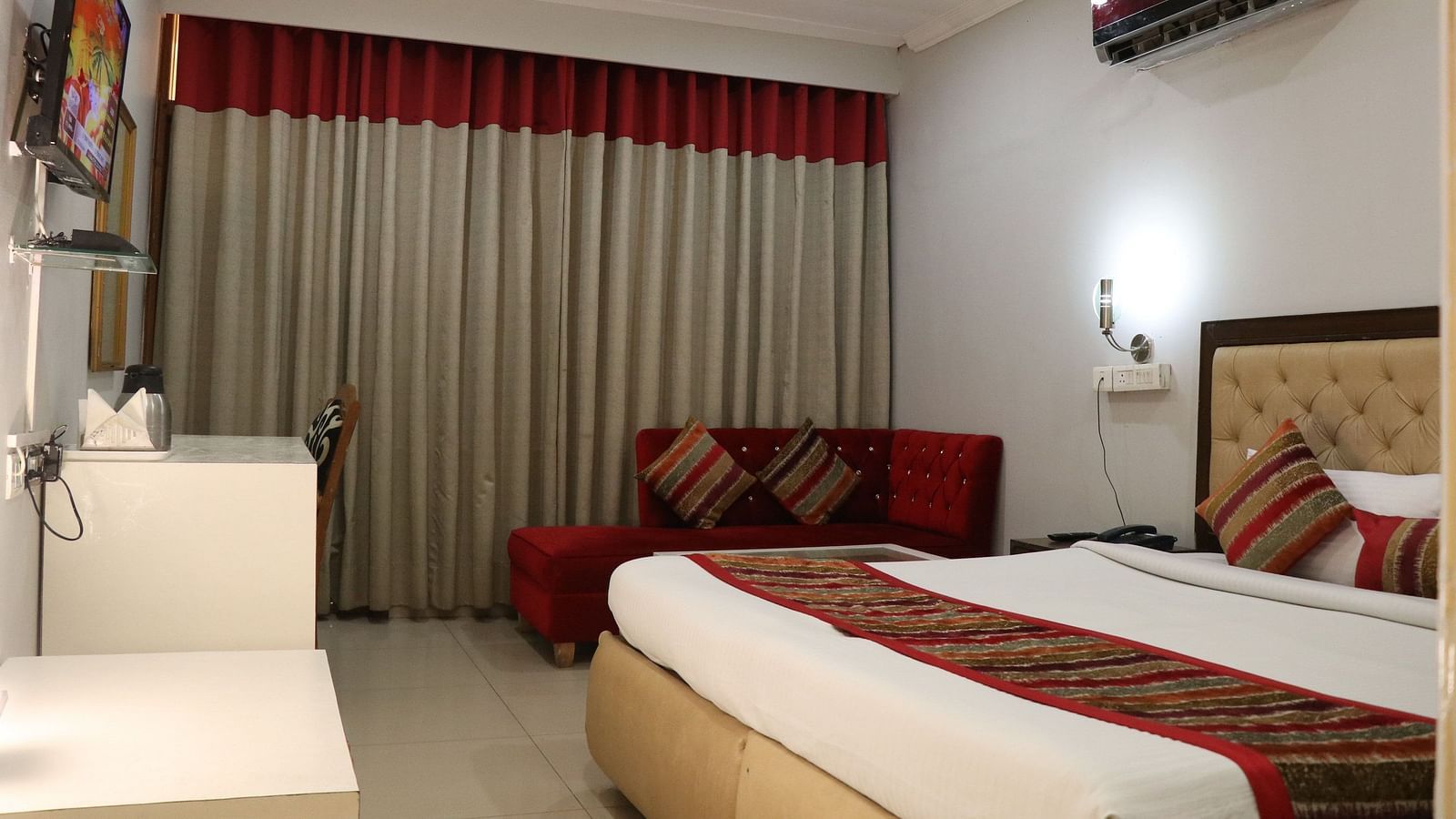 A hotel room with a plush bed featuring a patterned runner, a red sofa with striped pillows, and layered curtains, illuminated by soft lighting. - Hotel Diamond Plaza, Chandigarh.