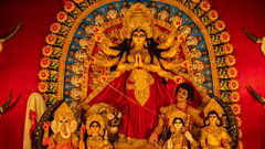 an idol of Goddess Durga with other deities below sitting next to her in a red backdrop