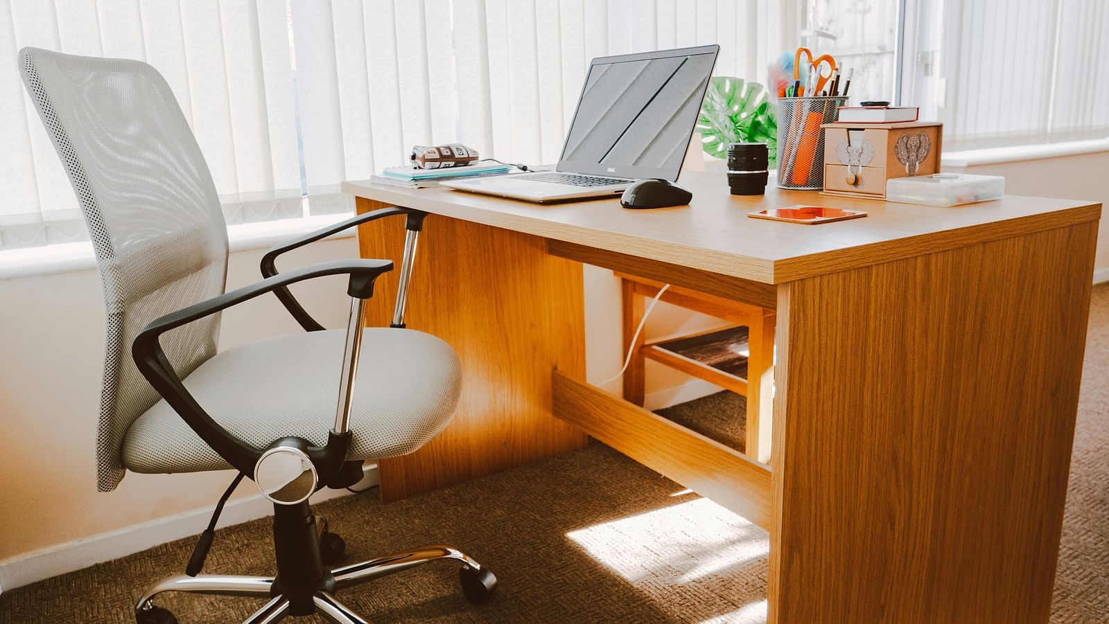 A office setup features a wooden desk with a laptop, mouse, stationery, and a grey ergonomic chair.