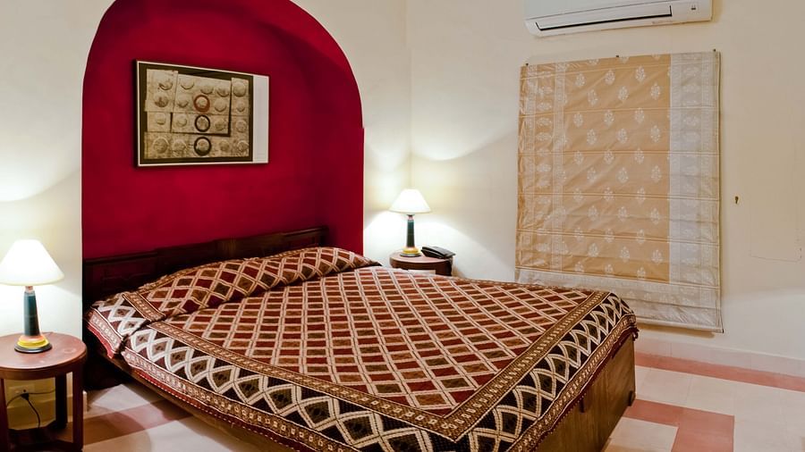 A bedroom with a patterned bedspread, a red arched alcove with a framed artwork, a decorative textile hanging on the wall, and an air conditioner, showcasing the room's unique design - Neemrana's Tijara Fort Palace.