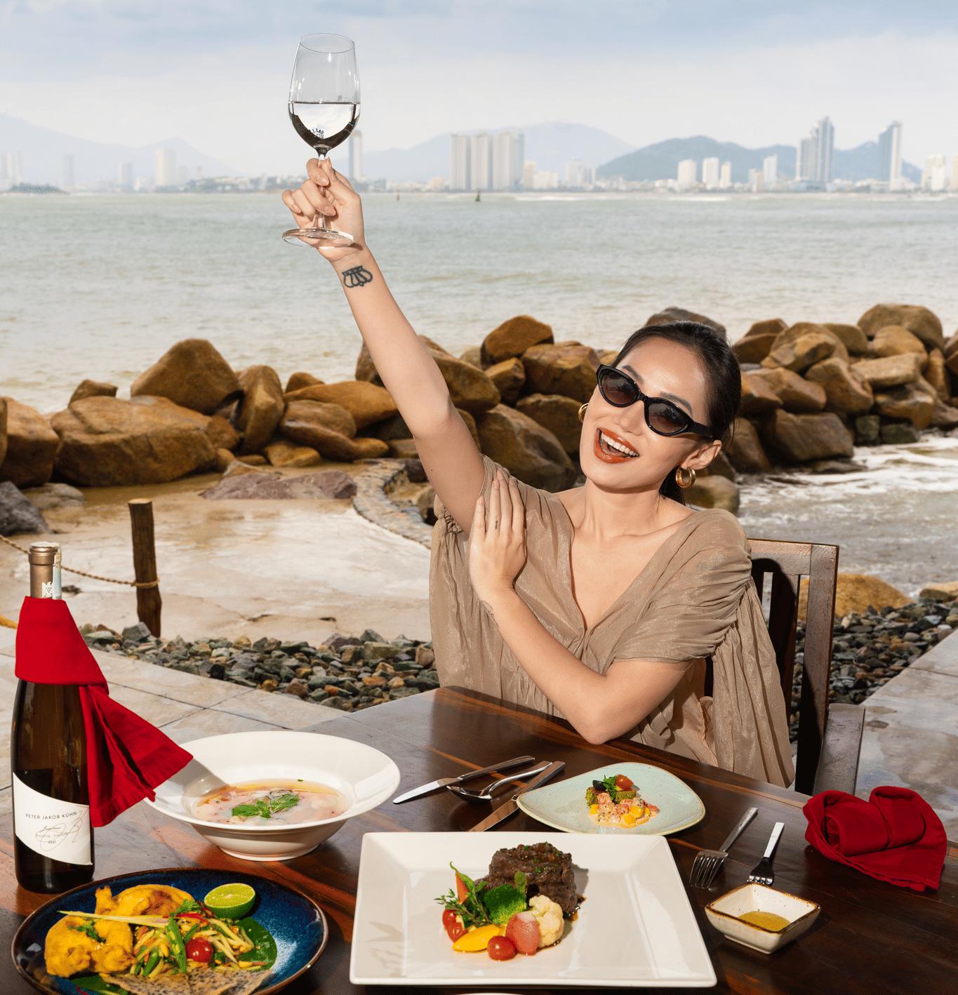 Woman sitting at a restaurant by the beachside featuring a dining table where several dishes are placed along with a liquor bottle.