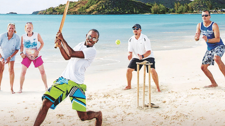 A group of people are playing cricket on a sandy beach with a lush green hill in the background.