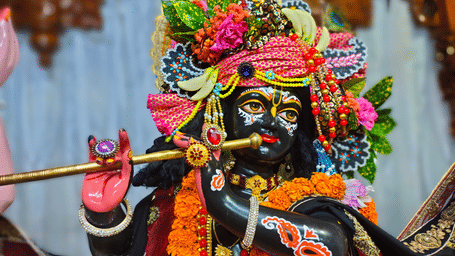 Detailed close up of a decorated Lord Krishna idol playing a flute, adorned with floral garlands, jewellery, traditional headwear, and ornamental decorations.