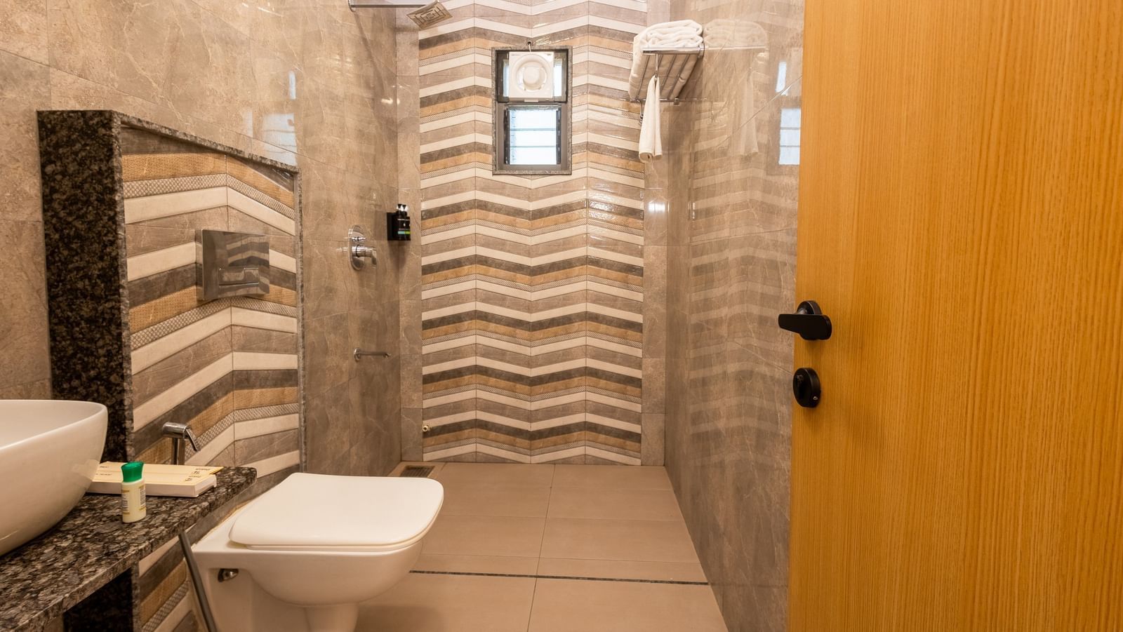 A modern bathroom at Daksh The Grand Gowardhan, Dwarka, showcasing its toilet, patterned wall tiles, and a wooden door.
